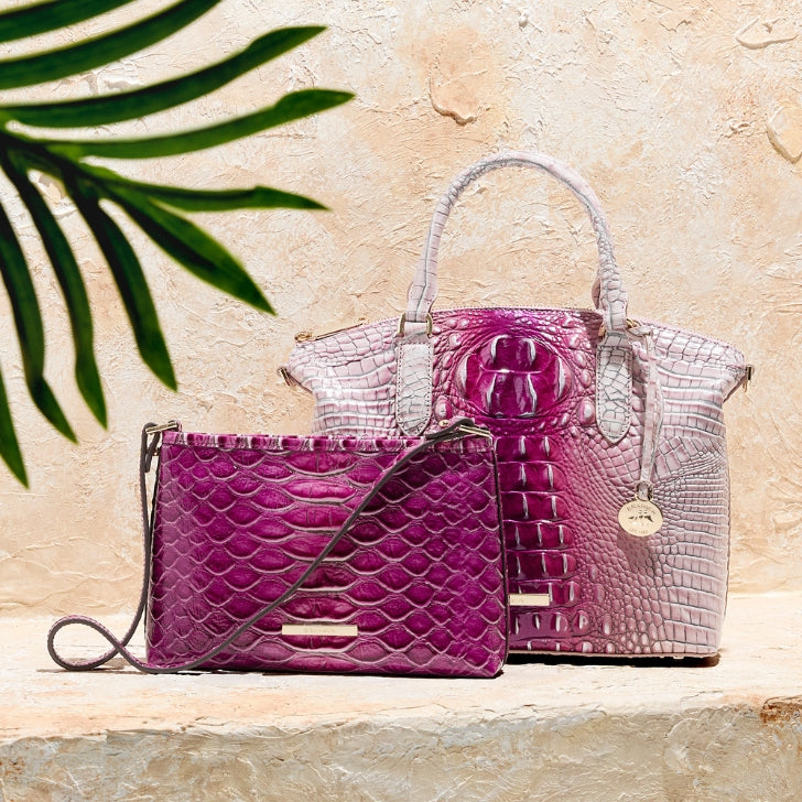 Coordinating Brahmin handbags displayed on a stone ledge, featuring a small magenta textured crossbody and a larger pink to plum ombré croc embossed tote beneath a palm frond.