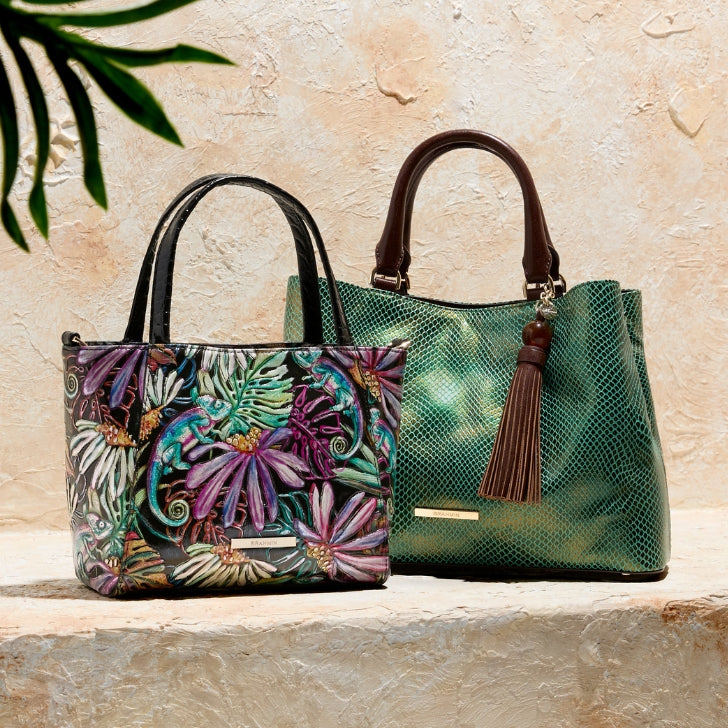 Two Brahmin tote handbags displayed on a stone ledge, including a multicolor tropical leaf print tote and a metallic green leather tote with a tassel beneath a palm frond.
