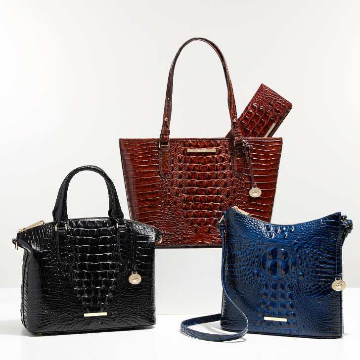 Three crocodile embossed leather handbags displayed against a white background, featuring a black satchel, rich brown tote, and deep blue crossbody bag with gold tone hardware and matching accessories.