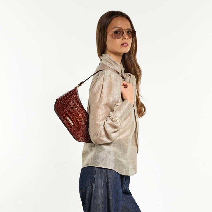 Model in a white blouse and blue jeans carrying a large glossy brown crocodile embossed shoulder bag against a white studio background.
