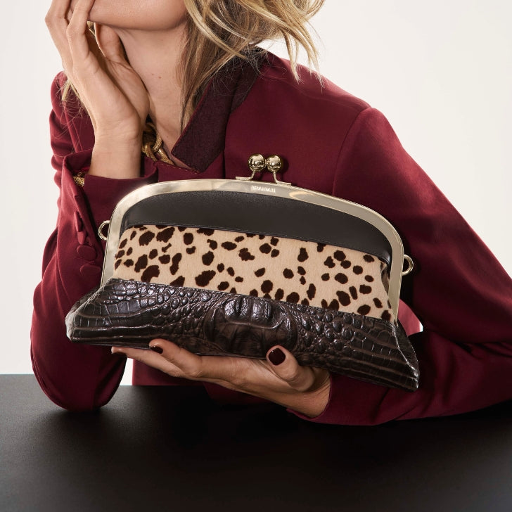 Model in a burgundy blazer leaning on a table while holding a structured Brahmin frame bag with leopard-print panel and chocolate brown croc-embossed leather trim.