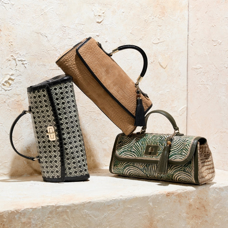 Three Brahmin satchel handbags styled on a stone ledge, with one tan textured bag positioned between a neutral geometric satchel and a green palm print satchel.